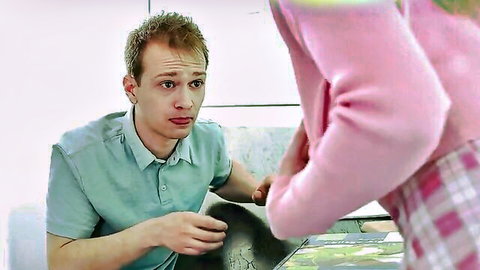 Young blonde in pink sweater and plaid skirt stands over surprised guy on couch