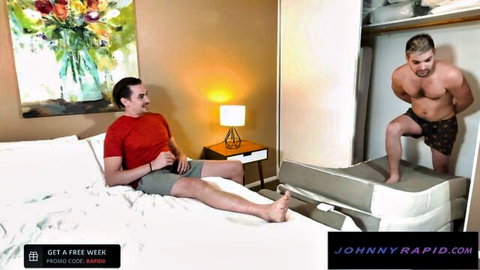 Muscular shirtless gay hunk in shorts stands by wardrobe as brunette stud in red shirt sits on bed teasing
