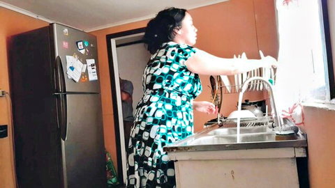 Mature Indian mom in tight patterned dress washing dishes at kitchen sink bending over