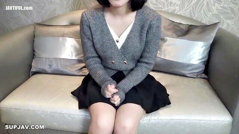 Cute Asian teen in gray cardigan and short black skirt sitting on couch with legs spread teasing
