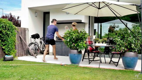 Couple enjoying outdoor BBQ with man grilling meat while blonde woman sits nearby on patio