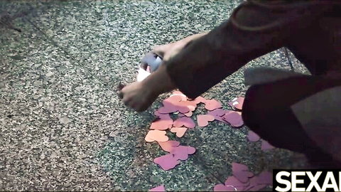 Clothed woman crouching with pink heart confetti on gravel in teasing outdoor scene