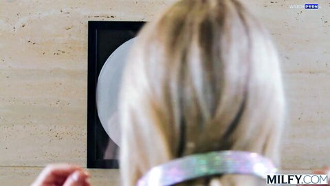 Rear view of long-haired blonde MILF reaching hands toward framed gloryhole on wall