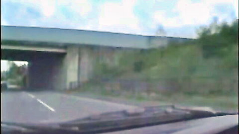 Car driving under highway bridge on road with bushes and cloudy sky.