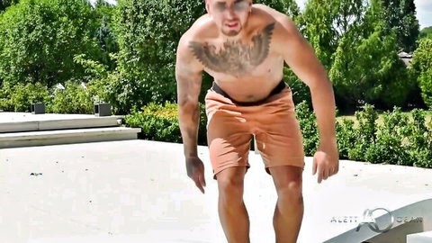 Muscular tattooed athletic guy flexing shirtless in tight orange shorts outdoors