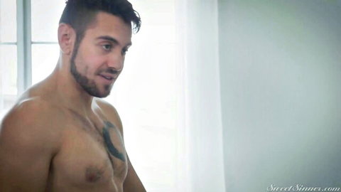 Muscular tattooed hunk with goatee stares intensely while shirtless in bedroom