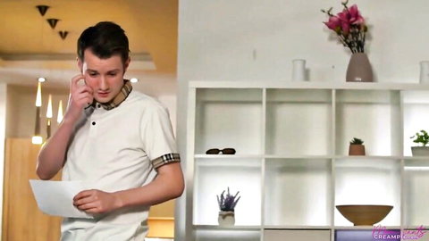 Young Caucasian man in white polo shirt talks on phone holding letter looking surprised in modern room