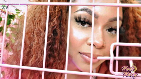 Seductive ebony babe with curly hair and glossy lips peeking through pink bars teasing for cock