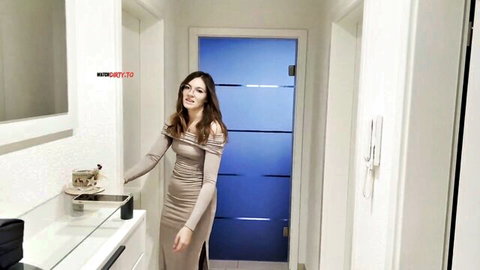 Brunette in tight beige dress teasing legs reaching for cup in bathroom hallway