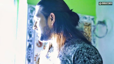 Bearded long-haired man in patterned shirt stands profile in green bathroom