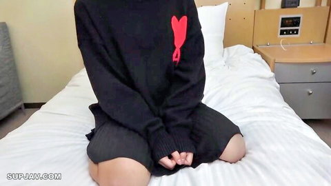 Cute Asian babe in oversized black sweater with red heart sits teasingly on hotel bed with thighs spread
