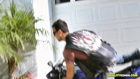 Biker stud in leather vest leaning over motorcycle in sunny garage