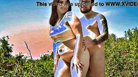 Curvy tattooed babe naked with shaved pussy embraces nude muscular guy with erect cock outdoors in grassy field at sunset