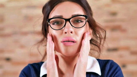 Shocked brunette teacher in glasses presses hands to surprised cheeks closeup