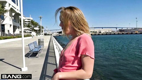 Blonde babe in low-cut pink dress teasing cleavage on sunny pier by ocean