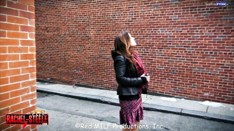 Rachel Steele, busty MILF in black leather jacket and red skirt against brick wall
