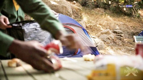 Dark skinned hands holding red apple over picnic table at campsite with blue tent and food