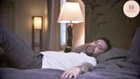 Tattooed bearded man lying asleep on bed in white t-shirt and gray pants