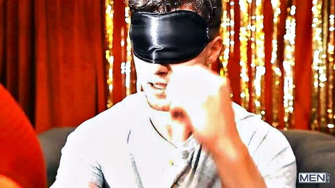 Blindfolded brunette muscular man grimacing with white cloth to mouth on couch in gay scene