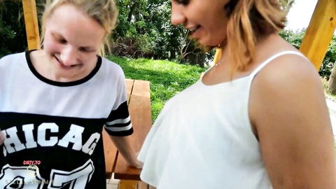 Blonde teen in Chicago shirt smiles teasingly with brunette in white top outdoors at picnic table