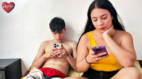 Shirtless young hunk in red briefs and curvy long-haired Asian babe in yellow top check phones on bed