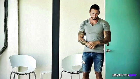 Tattooed hunk Logan Cross in gray tee and ripped shorts by door