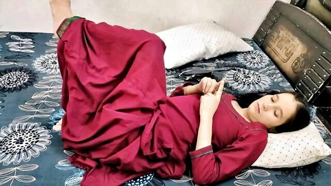 Sexy Indian wife in red salwar kameez lying asleep on bed with legs up holding phone