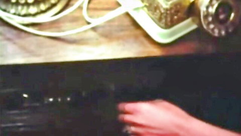 Woman's hand with red nails reaches for VCR buttons beside gold rotary phone on wooden nightstand
