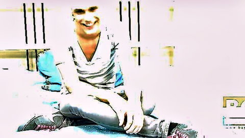 Smiling blonde guy sitting cross-legged in tight jeans and v-neck shirt in bedroom