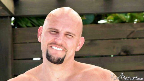Shirtless bald hunk with goatee smiles seductively outdoors by pool