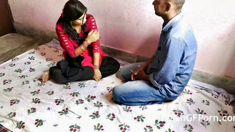 Indian girlfriend in red top and black salwar sits facing boyfriend on floral bed