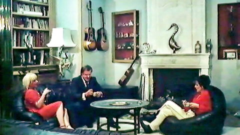 Blonde and brunette women in red outfits with suited man in cozy living room