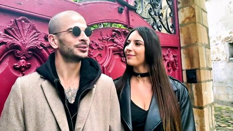 Bald hunk in sunglasses smirks with busty brunette in choker and leather jacket posing outdoors