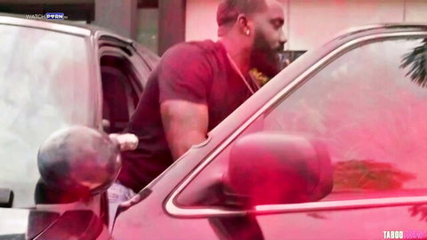 Muscular black stud in red shirt leans into red car door in taboo outdoor scene