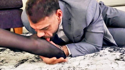 Suited man passionately licks woman's black nylon-clad foot on the floor