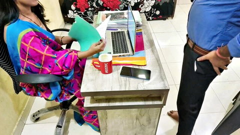 Indian boss stands behind sexy saree-clad secretary at office desk holding papers