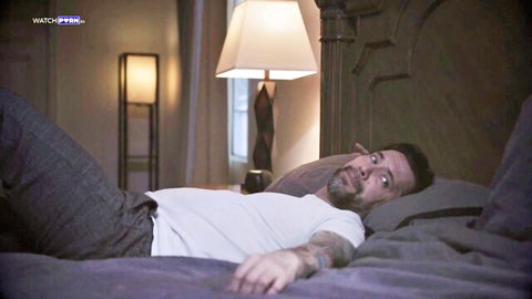 Tattooed bearded man lying on bed in gray pants and white t-shirt