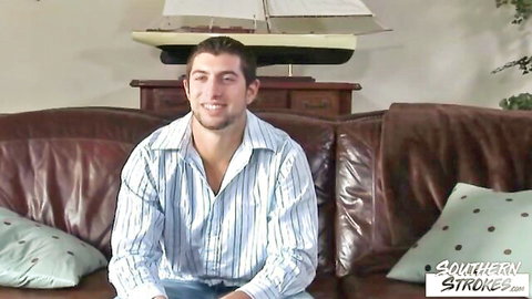 Handsome young athletic guy smiles in open striped shirt on couch during gay interview