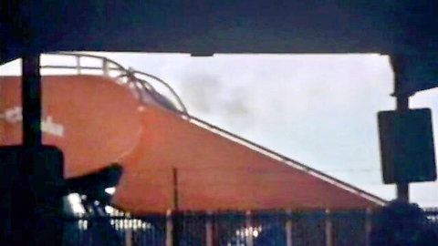 Large orange curved slide structure with black railing and fence in outdoor playground setting
