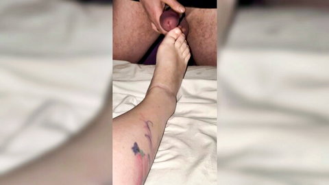 Woman's tattooed bare foot teasing man's hard erect cock held by his hand in footjob.