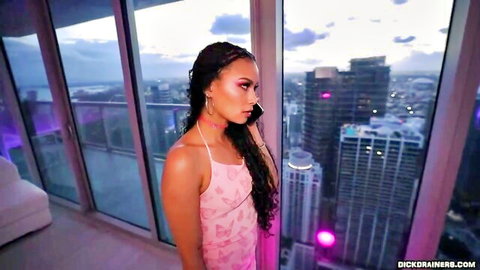 Tanned petite mixed brunette in tight pink dress on phone by Miami penthouse window