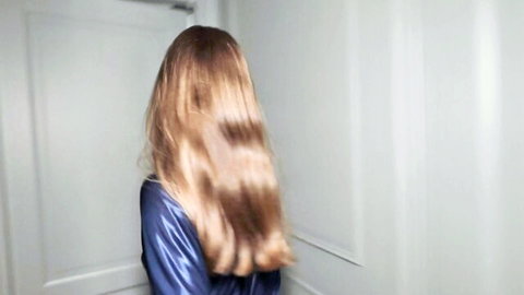 Blonde woman with long wavy hair in shiny blue satin robe viewed from behind teasing seductively.