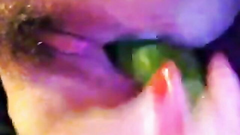 Closeup of woman's half-closed eye teased by thick green dildo held by red-nailed fingers