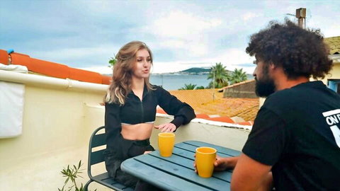 Blonde teen in black crop top chats teasingly with curly-haired black guy sipping yellow mugs on sunny terrace