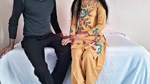 South Asian couple in traditional embroidered outfits sitting closely on bed teasing