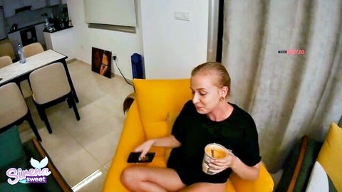 Skinny blonde teen sitting legs spread on yellow couch holding phone and cup in black outfit