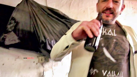 Bearded man in Valhalla t-shirt drinks beer casually with trash bag behind