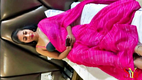 Sexy Indian woman in sheer pink saree revealing black bra cleavage lying seductively on hotel bed