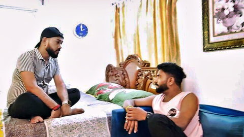 Two handsome Indian men in tank top and shirt chatting intimately on bed and chair in bedroom