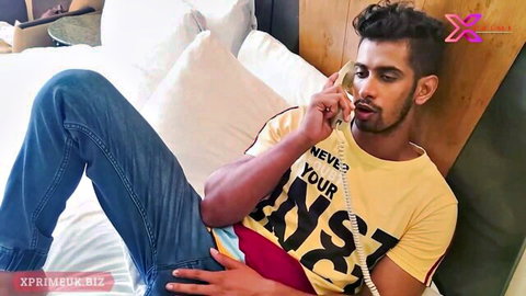 Desi teen guy lounging on bed in yellow shirt and jeans, making a flirty call